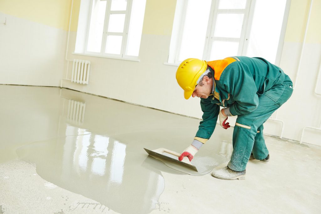 A man in a hard hat and green jumpsuit painting a floor
