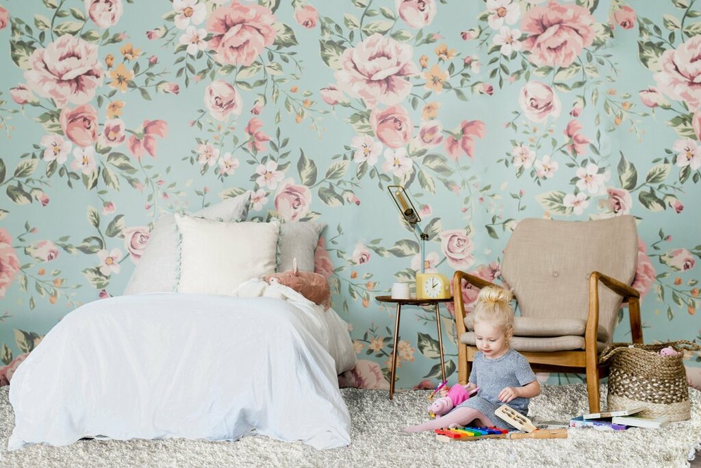 A little girl sitting on the floor in front of a floral wallpaper
